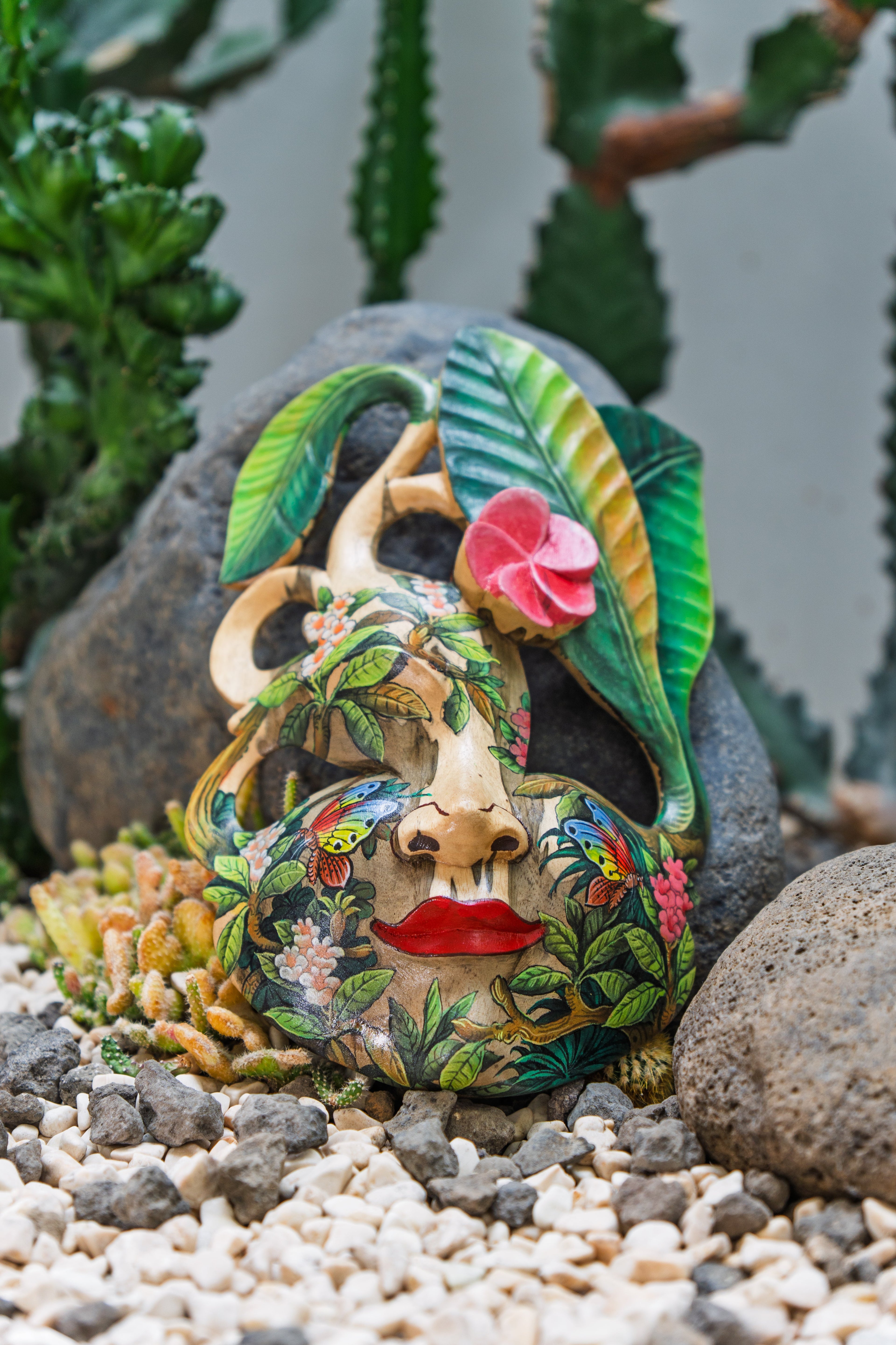 a colourful wooden facemark laying on the bright gravel ground. masked is full of flower patterns and the top is shaped like a leaf with a blossom over it. 